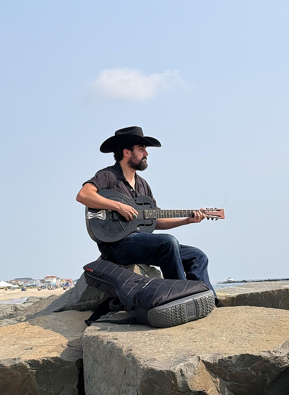 Bearded man wearing black cowboy hat playing acoustic guitar while sitting on rocks overlooking a desert landscape under a clear blue sky.