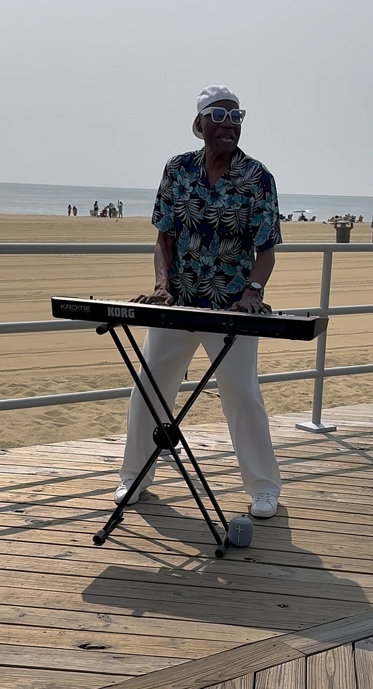 Man playing keyboard on a wooden boardwalk at the beach wearing sunglasses and a tropical print shirt.