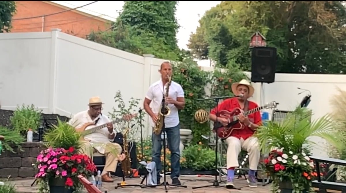 Three musicians perform outdoors on a stage, surrounded by colorful flowers and potted plants in a residential garden setting.