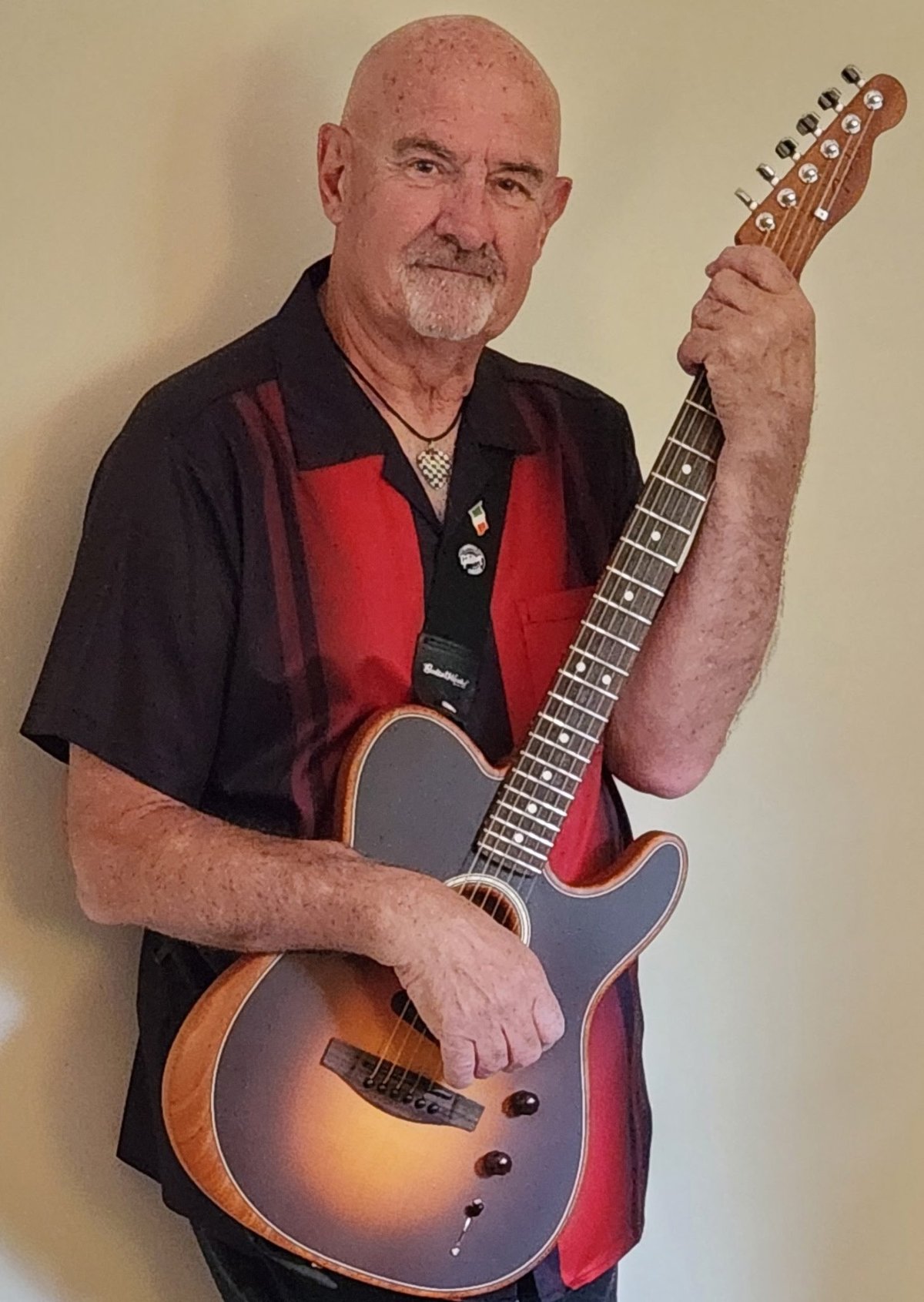 Bald man with a mustache holding a sunburst acoustic guitar, wearing a black and red shirt.